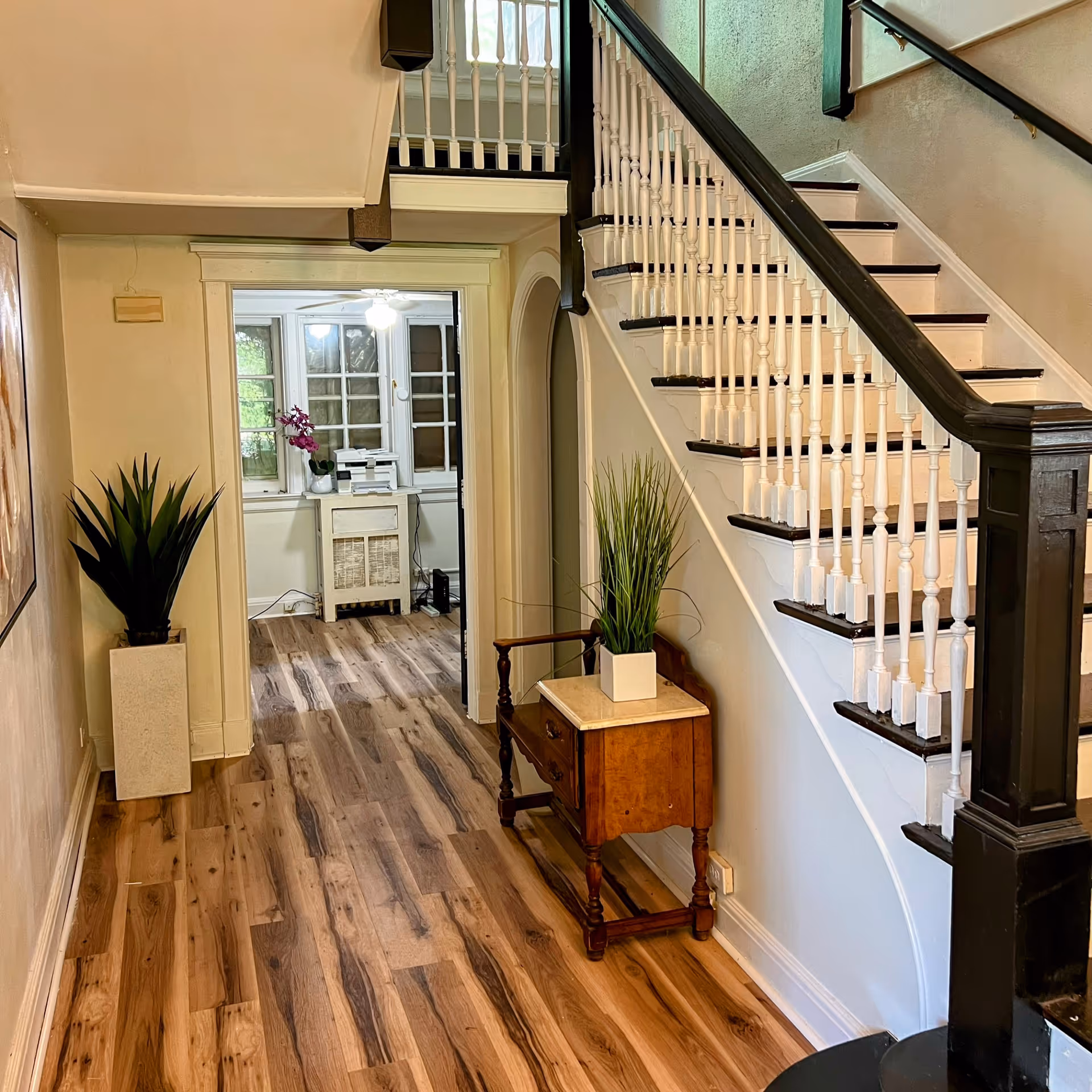 Interior view of a senior living facility hallway with wooden flooring, a staircase with white spindles and dark handrails, a small wooden table with a potted plant, and another potted plant in a tall rectangular planter. A room with windows and a ceiling fan is visible at the end of the hallway.