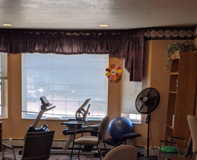 A small exercise room with two stationary bikes, a blue exercise ball, several chairs, a standing fan, and a wooden shelving unit with some items on it. The room has large windows with blinds and purple valances, and a colorful flower decoration on the wall.