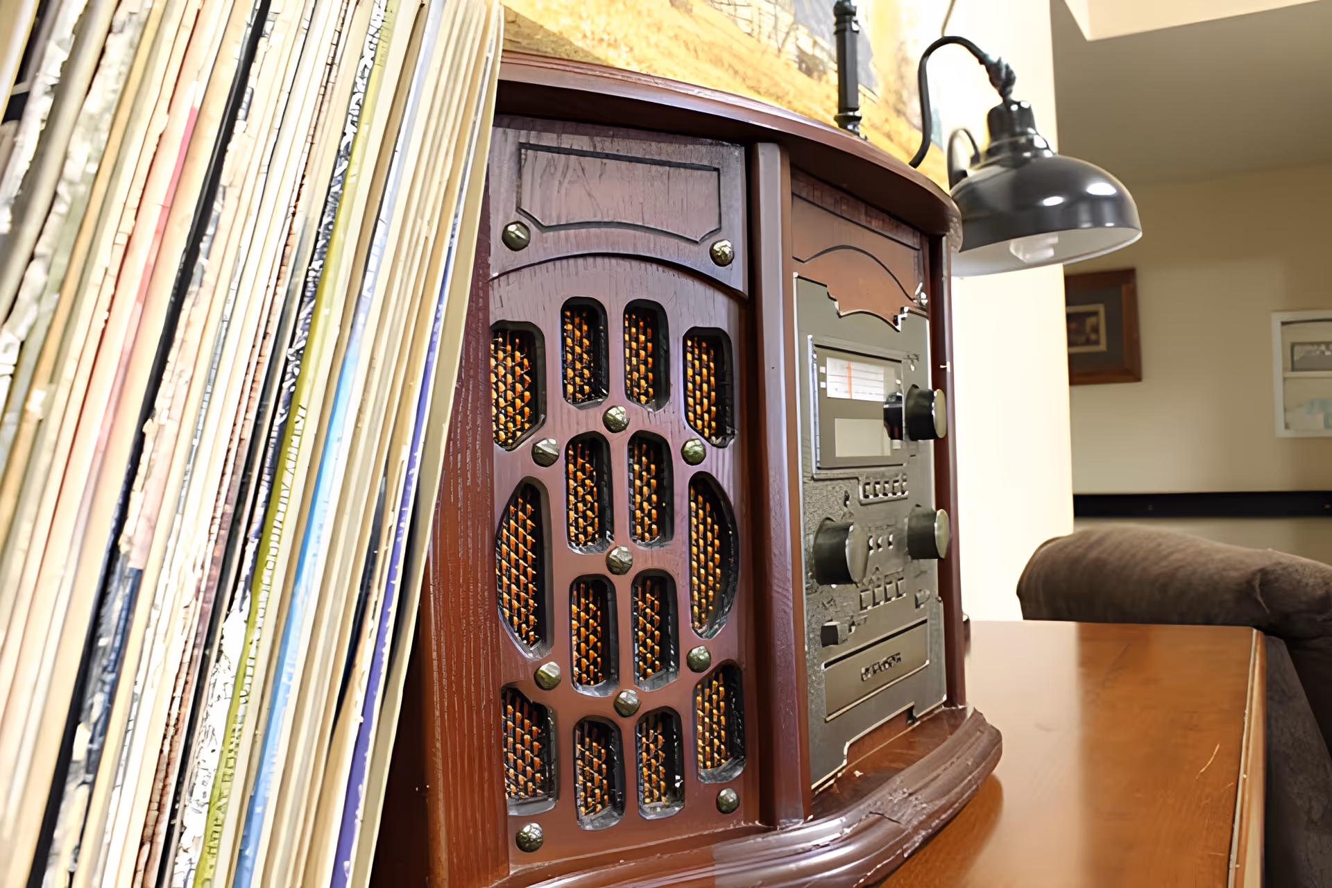 Close-up view of a vintage-style wooden radio on a wooden table with a stack of vinyl records leaning against it, a black desk lamp attached to the table, and a brown couch in the background in a living room setting.