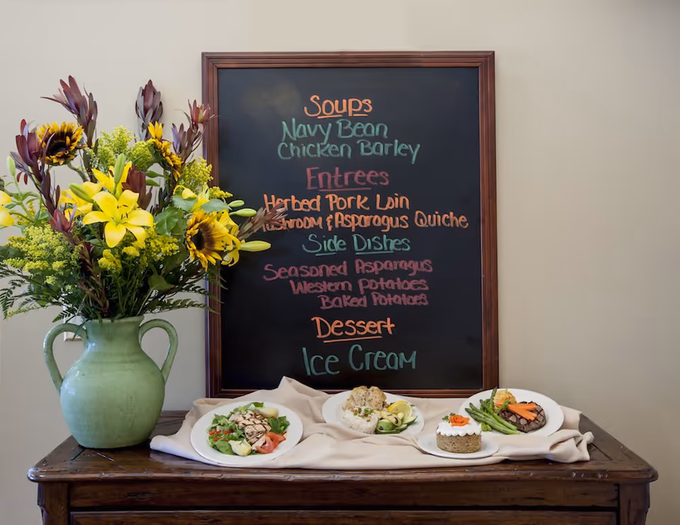 A wooden table with a green vase holding a bouquet of yellow lilies, sunflowers, and other flowers. Behind the vase is a chalkboard menu listing soups, entrees, side dishes, and dessert. On the table in front of the chalkboard are four plates of food including a salad, a dish with rice and vegetables, a dessert with frosting and a carrot decoration, and a plate with asparagus and carrots.