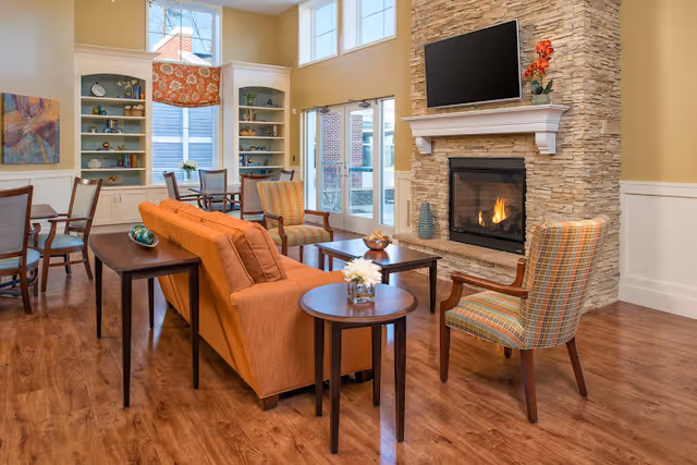 Bright communal living room with seating, a stone fireplace, wall-mounted TV, and large windows.