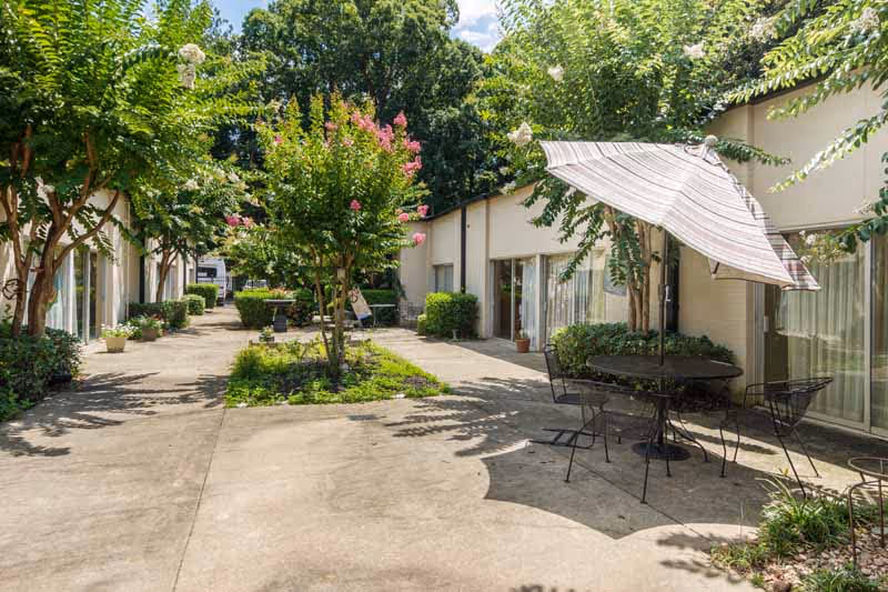 Sunlit courtyard between single-story buildings with trees, potted plants, and a patio table with umbrella.