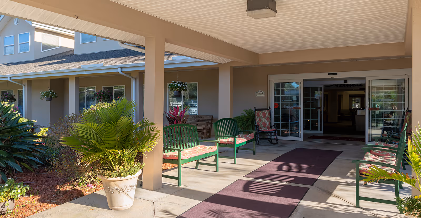 Covered entrance with green benches, potted plants, and sliding glass doors leading into a senior living community.