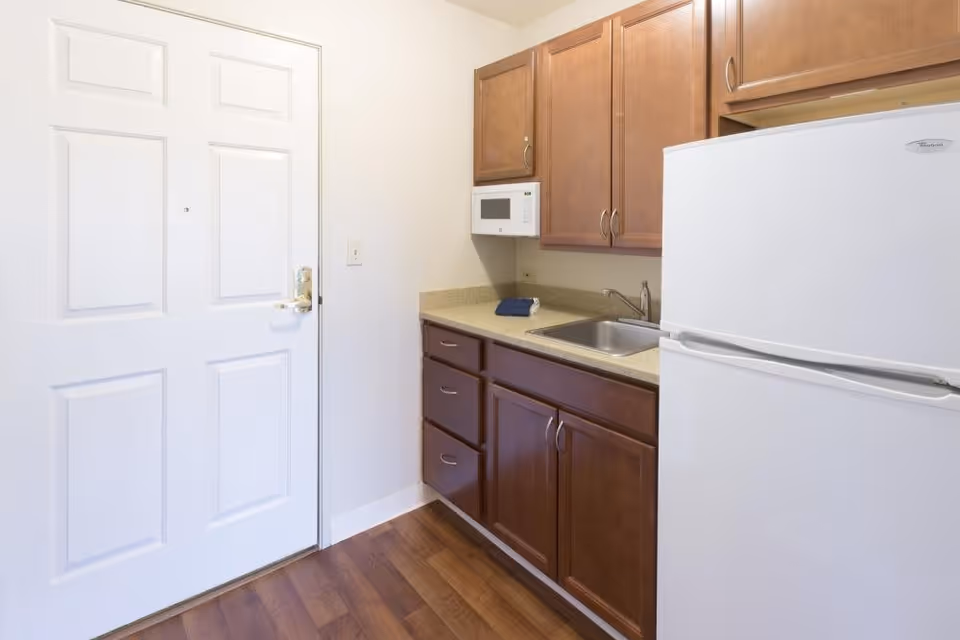 Small kitchen area with wooden cabinets, a countertop with a sink, a white microwave mounted above the counter, and a white refrigerator. A white door is visible to the left, and the floor has wood-style flooring.