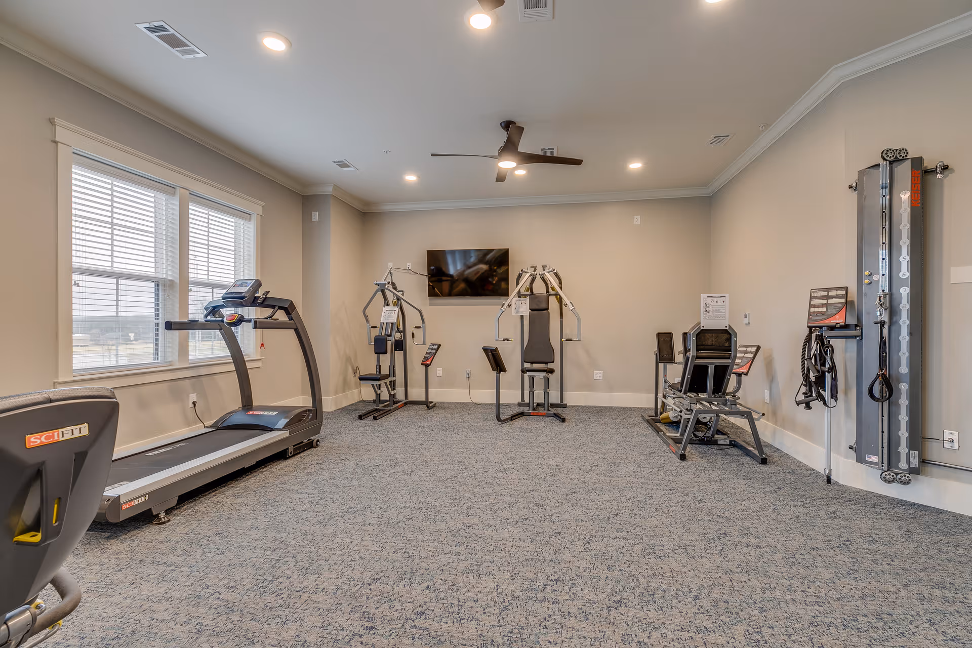 A fitness room with exercise equipment including a treadmill, weight machines, and a wall-mounted TV. The room has beige walls, carpeted floor, recessed ceiling lights, a ceiling fan, and large windows letting in natural light.