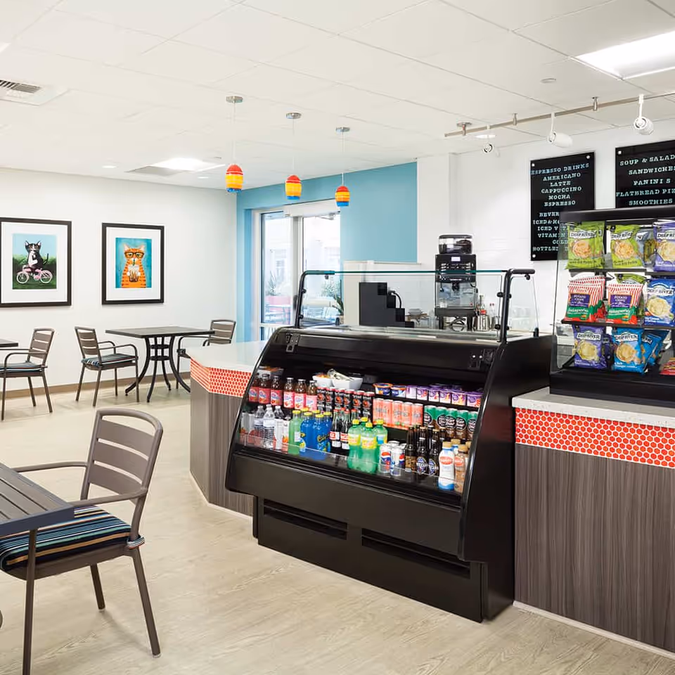 Interior view of a small café or snack bar area in Bayview Retirement Community featuring a refrigerated display case stocked with beverages and snacks. There are tables and chairs for seating, colorful pendant lights hanging from the ceiling, and framed pictures of animals on the wall.
