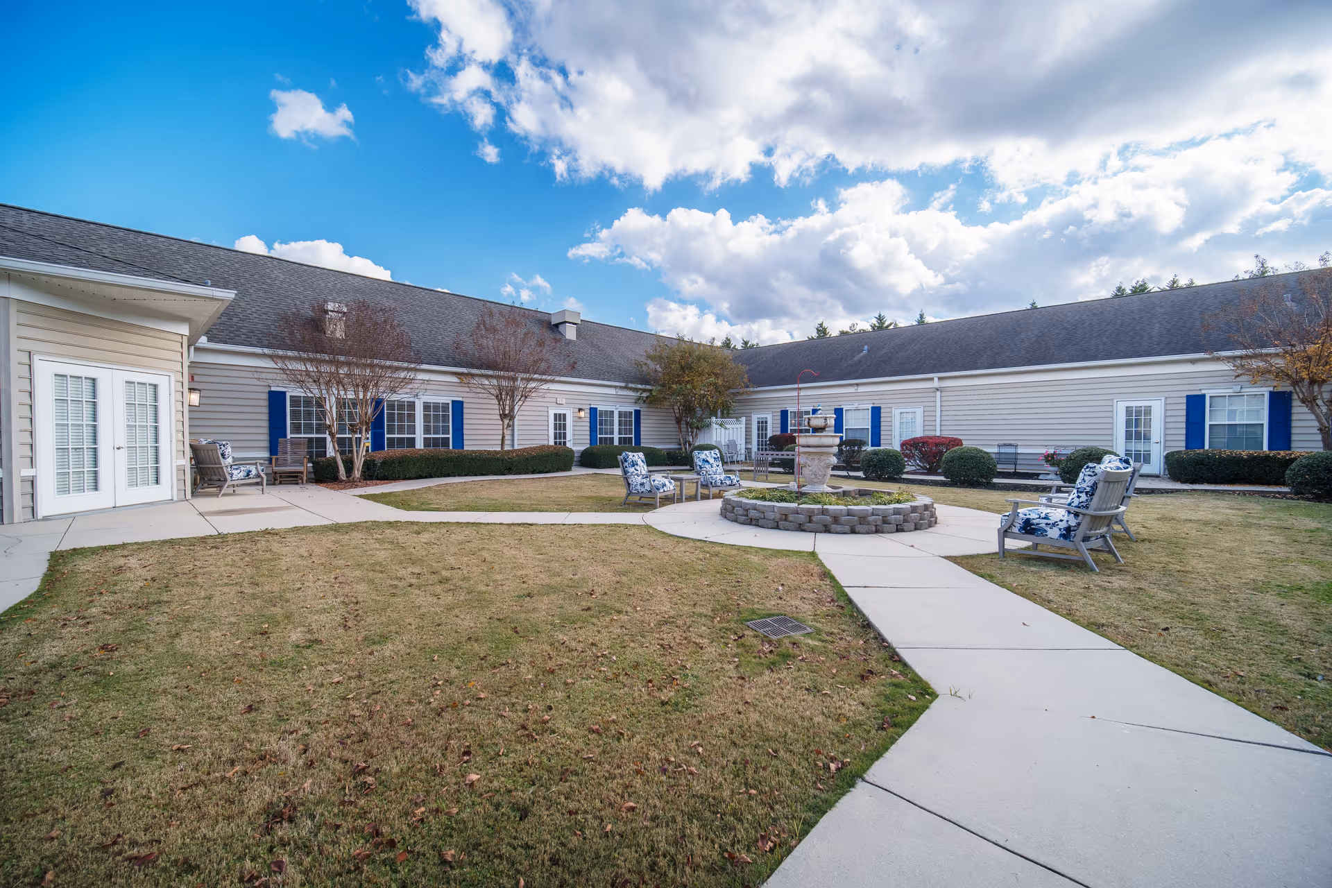 Outdoor courtyard area of a senior living facility with a circular stone fountain in the center surrounded by cushioned chairs. The courtyard is enclosed by a single-story building with beige siding, blue shutters, and multiple windows and doors. The sky is partly cloudy with patches of blue.