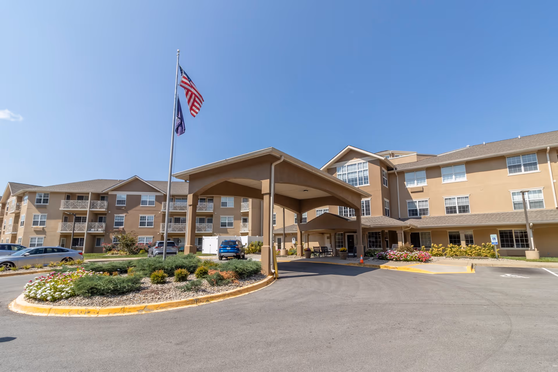 Exterior view of a multi-story senior living facility with a covered entrance driveway, landscaped flower beds, and two flagpoles displaying the American flag and another flag under a clear blue sky.