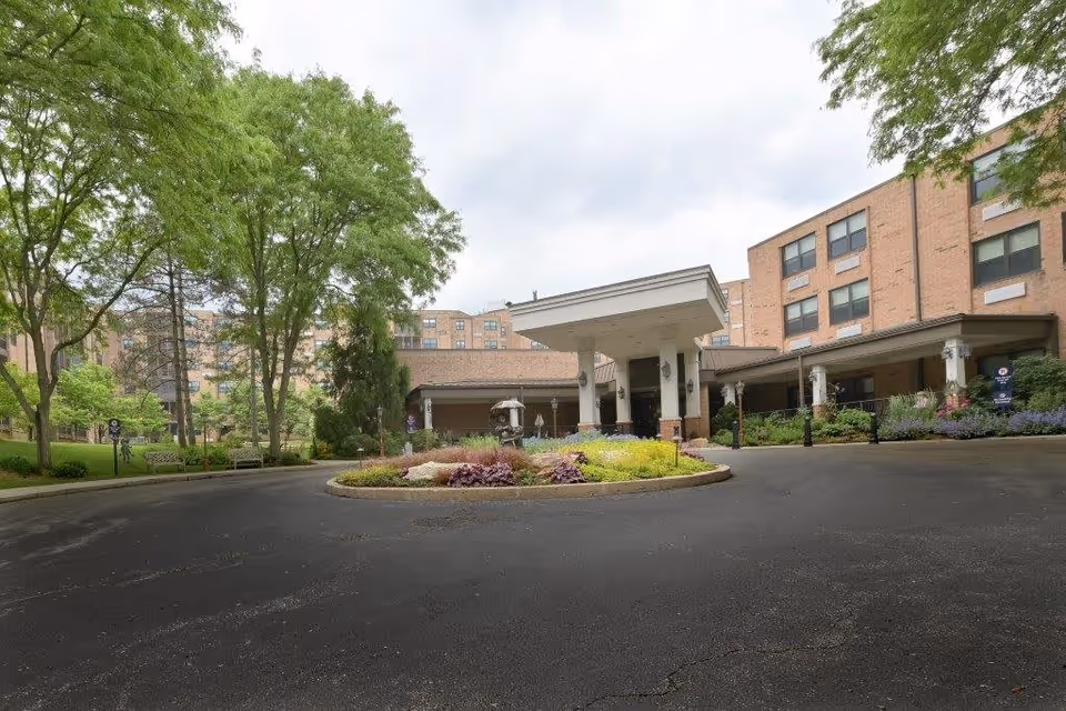 Front exterior view of The Lafayette facility showing a circular driveway with a landscaped island in the center, surrounded by trees and a multi-story brick building with large windows and a covered entrance.