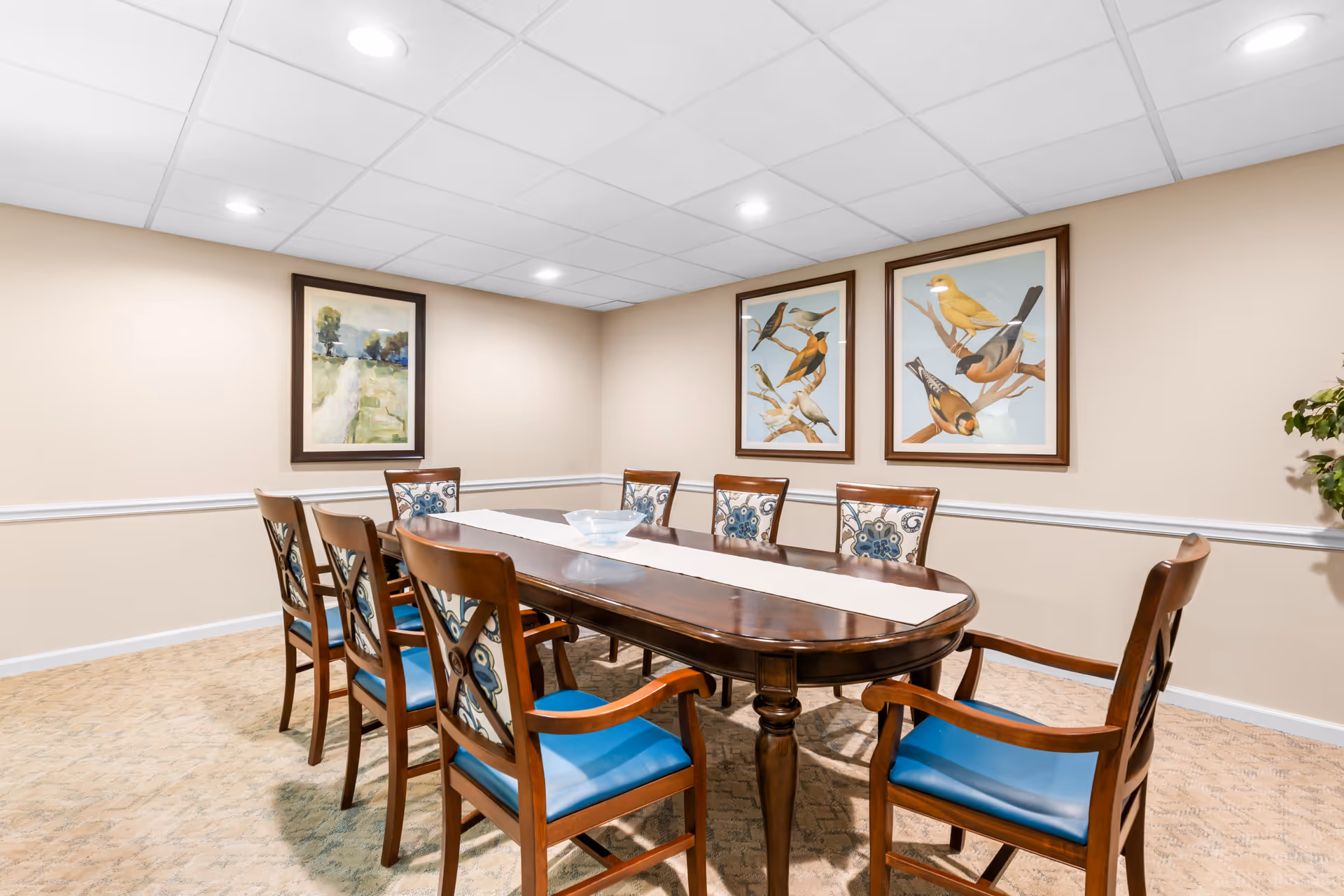 A dining room with a long wooden table surrounded by eight wooden chairs with blue cushioned seats and patterned backs. The walls are beige with white trim, and three framed artworks hang on the walls, including two pictures of birds and one landscape painting. The ceiling has recessed lighting.