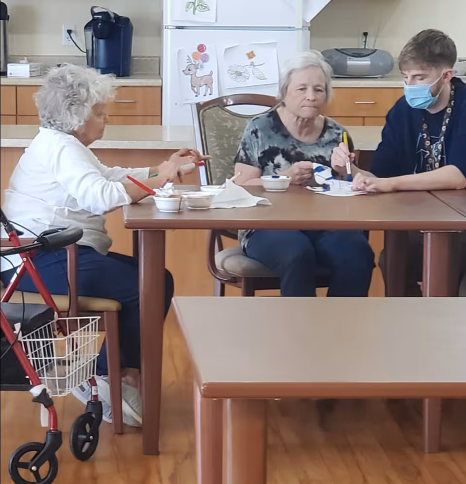Two elderly women and a masked staff member sit at a table in a communal dining/activity room doing crafts and eating.