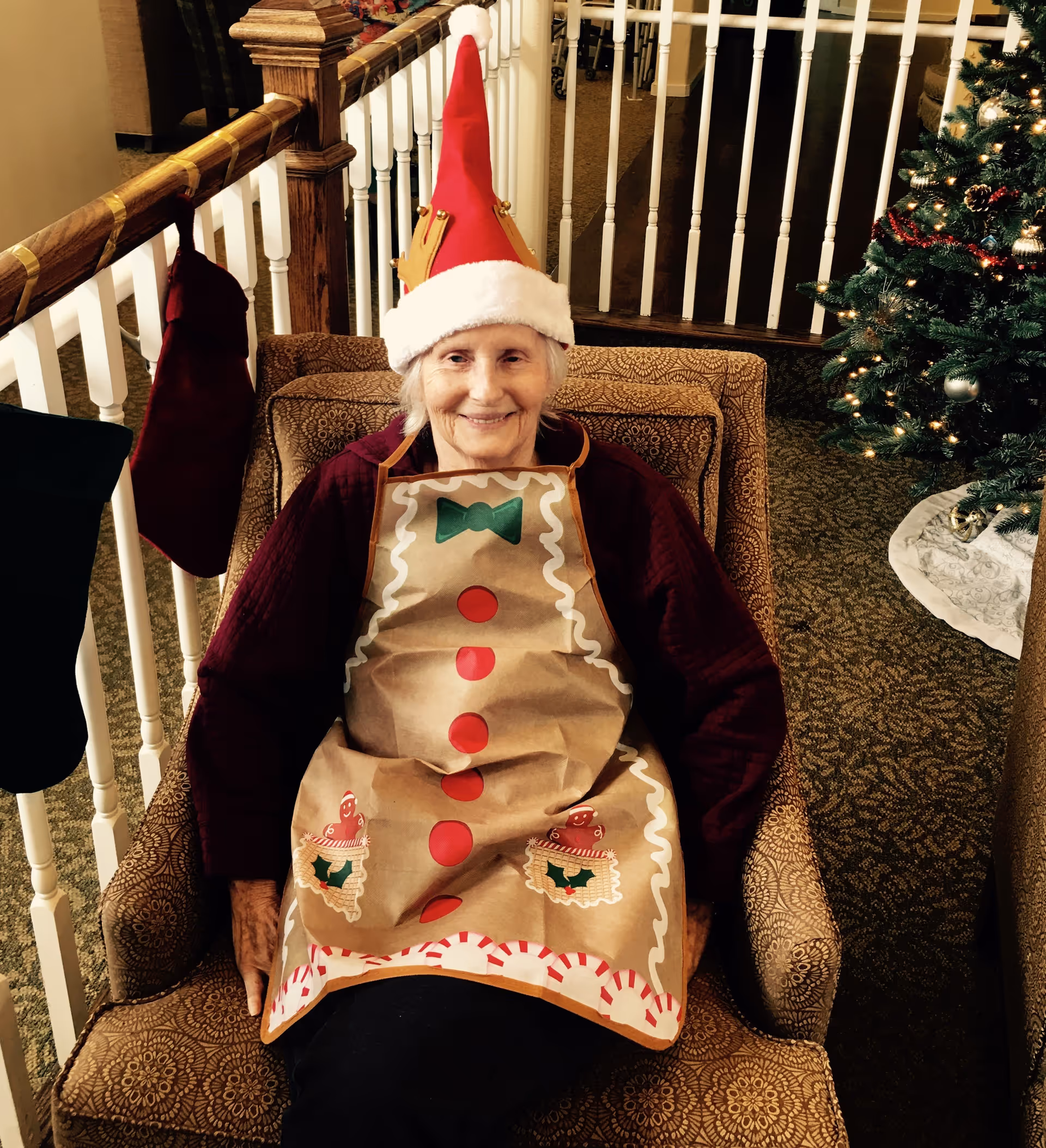 An elderly woman sitting in a patterned armchair wearing a festive red Santa hat with a gold crown and a gingerbread-themed apron. Behind her is a white railing with Christmas stockings hanging on it and a decorated Christmas tree with lights and ornaments to the right.