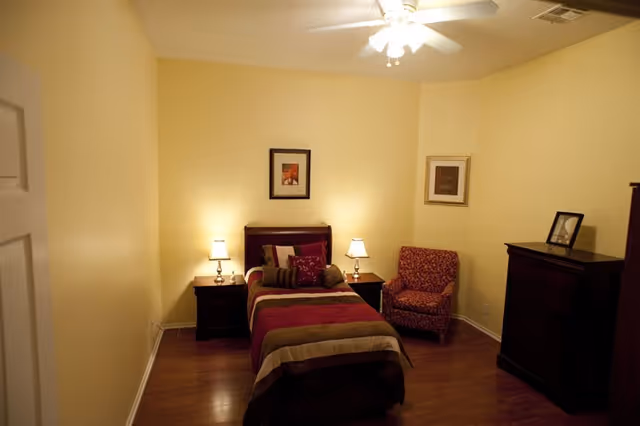 A cozy bedroom with a single bed featuring a striped bedspread in shades of brown, beige, and burgundy. There are two bedside tables with lamps on each side of the bed. A patterned armchair is placed in the corner next to a dark wooden dresser with a framed photo on top. The walls are painted light yellow, and two framed pictures hang above the bed and on the adjacent wall. The room has wooden flooring and a ceiling fan with lights.