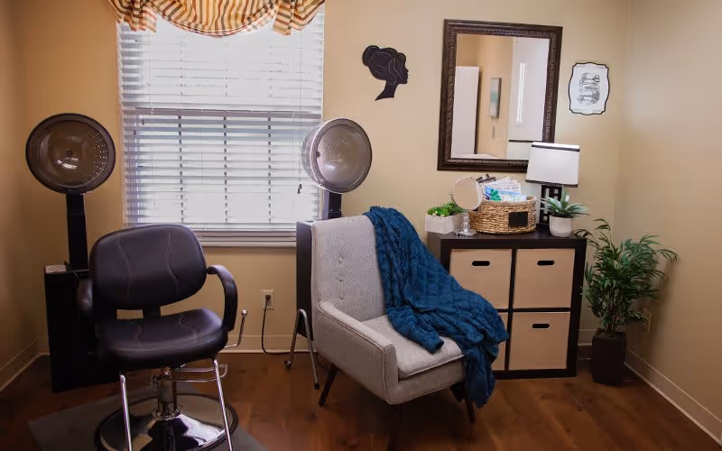 A cozy room with a black salon chair and two hair dryer stations near a window with blinds and a striped valance. There is a gray armchair with a blue knitted blanket draped over it. Next to the armchair is a black storage unit with four beige fabric drawers, topped with a basket, a small mirror, a lamp, and a potted plant. A large framed mirror and wall decorations hang on the beige walls. The floor is wooden.