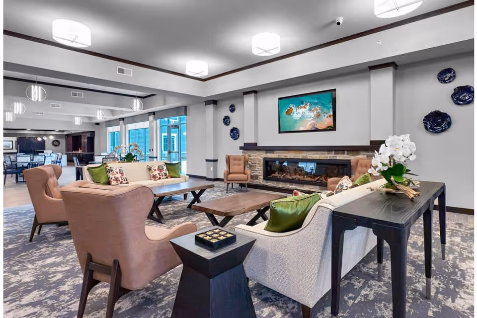 A spacious and modern living room area in a senior living facility featuring comfortable armchairs and sofas with green and floral cushions arranged around wooden coffee tables. A long black console table with a white orchid plant is behind one sofa. The room has a stone fireplace with a colorful abstract painting above it and decorative blue plates on the wall. Large windows allow natural light to fill the space, and a dining area is visible in the background.