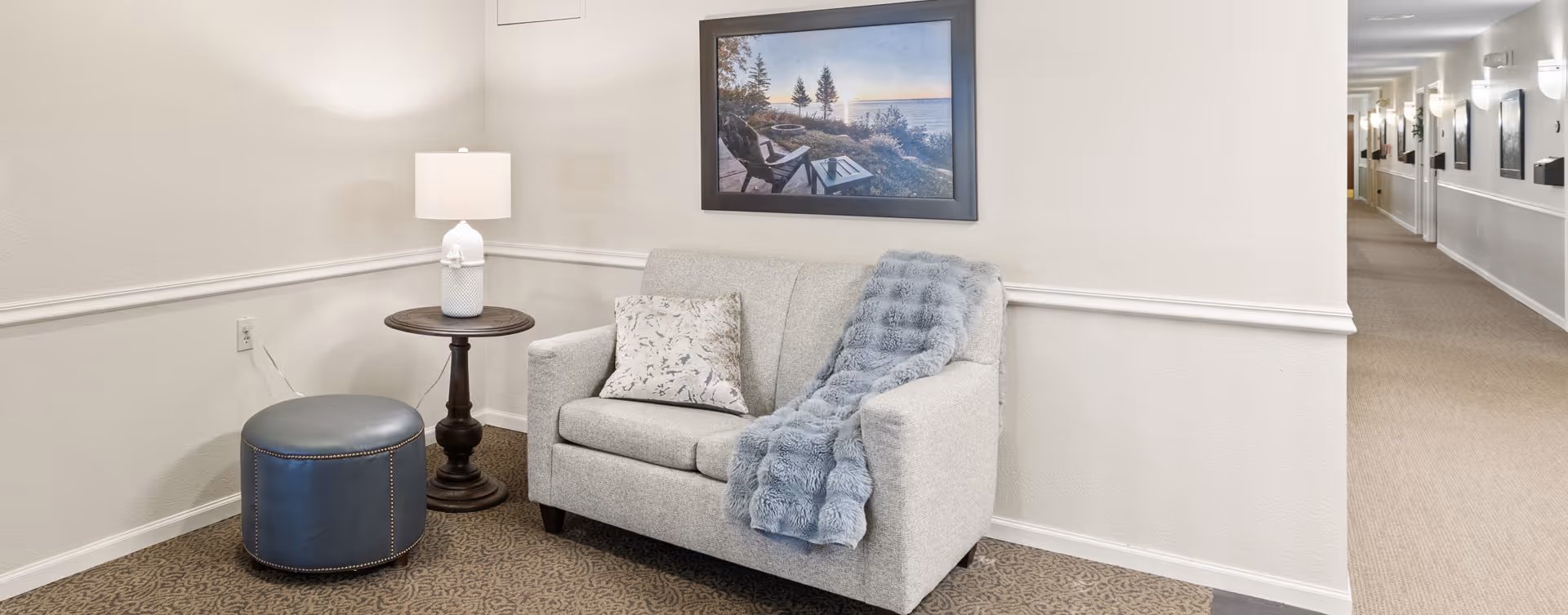 A cozy seating area with a light gray loveseat adorned with a patterned pillow and a blue throw blanket. Next to the loveseat is a round wooden side table with a white lamp on it, and a blue ottoman. A framed picture of a lakeside scene hangs on the wall above the loveseat. The area is carpeted and adjacent to a hallway with several wall-mounted lights and mirrors.