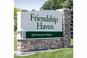 Outdoor stone sign with white and green panels displaying the name Friendship Haven and the address 420 Kenyon Road, surrounded by grass and trees in the background.