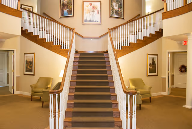 Symmetrical two-story foyer with a central carpeted staircase that splits into two wooden balustrade flights, flanked by armchairs, lamps, and framed artwork.