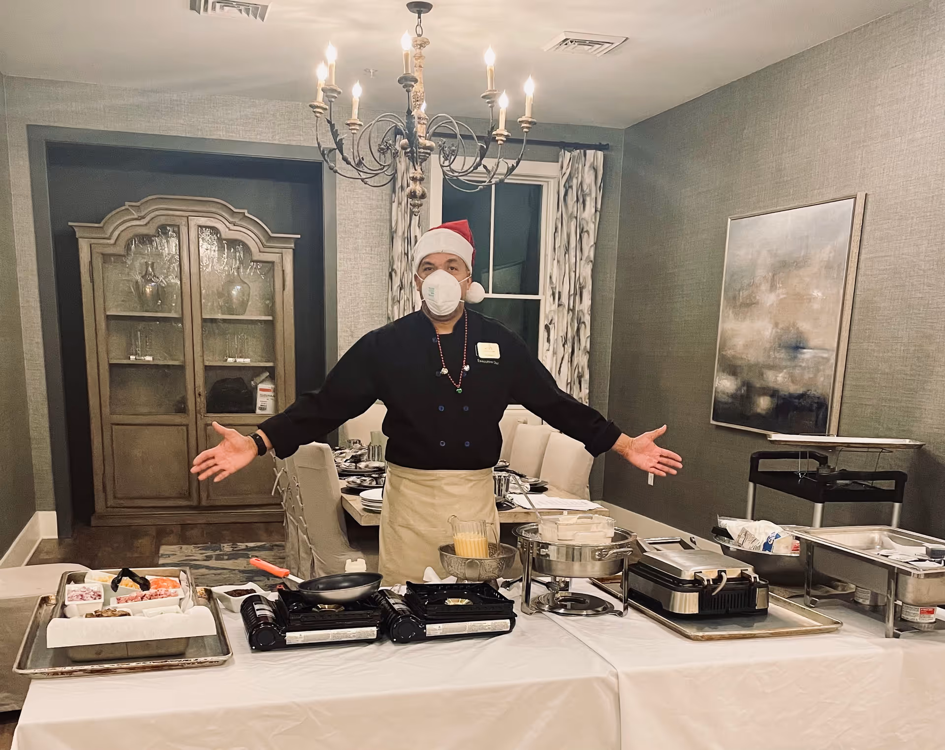 A person wearing a Santa hat, face mask, and black chef's jacket stands behind a table set up with cooking equipment and ingredients in a dining room. The room features a chandelier, a wooden cabinet with glassware, a window with patterned curtains, and a painting on the wall.