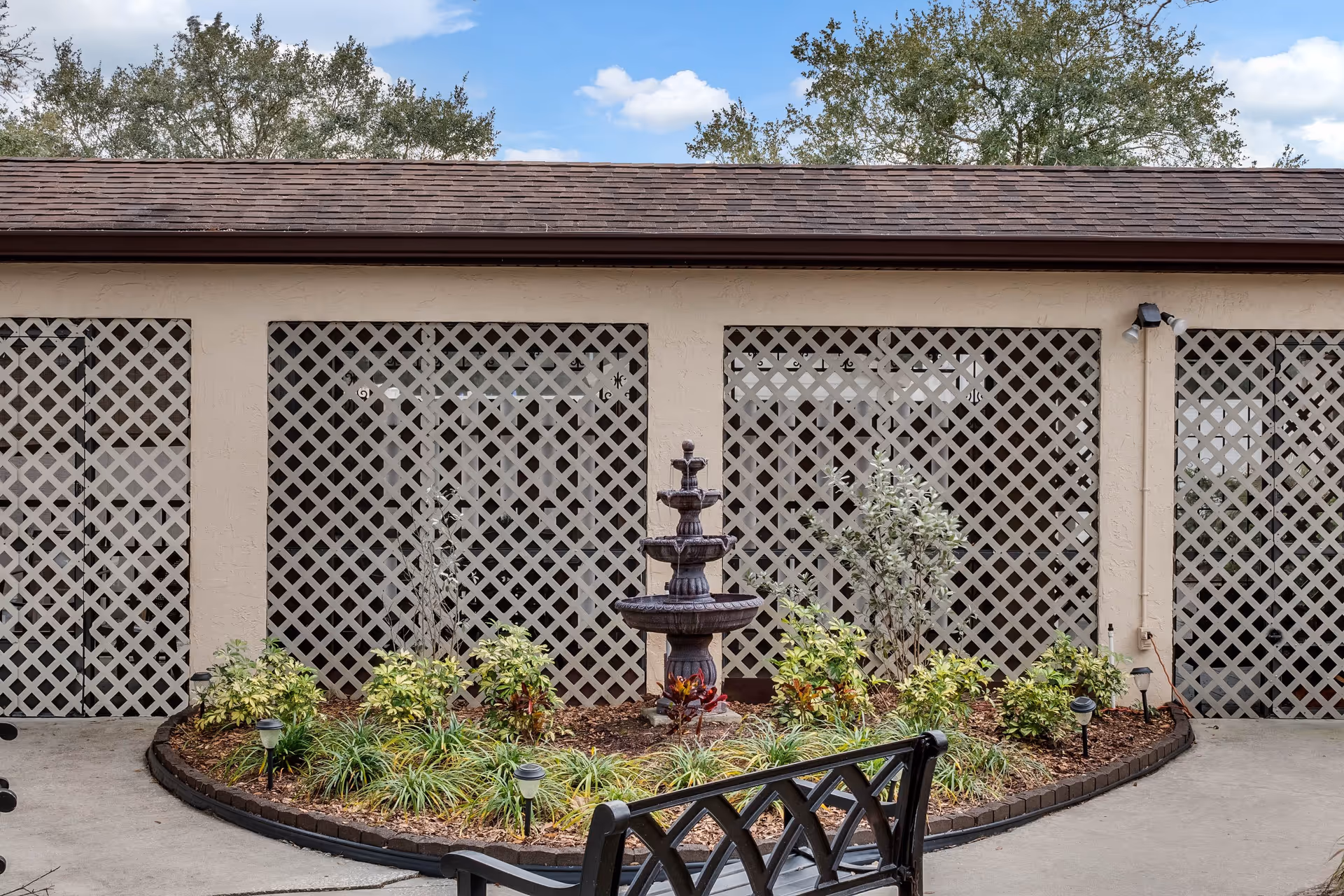 Outdoor courtyard with a tiered fountain, landscaped flowerbed, lattice-covered wall, and a bench.
