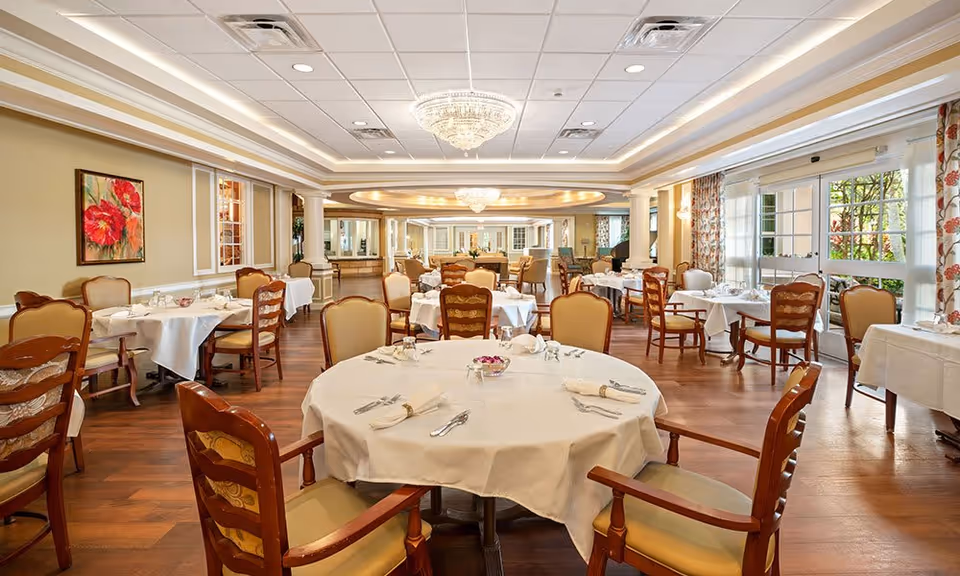 A spacious and elegant dining room with round and rectangular tables covered with white tablecloths, set with silverware and napkins. The room features wooden chairs with cushioned seats, large windows with floral curtains allowing natural light, chandeliers hanging from the ceiling, and a painting of red flowers on the wall.