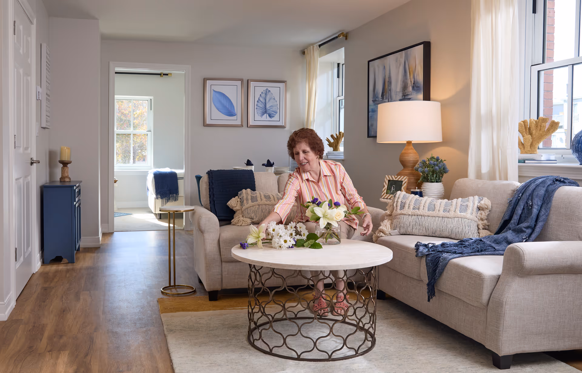 A cozy living room with beige sofas adorned with textured pillows and a blue throw blanket. A woman with short curly hair wearing a striped shirt is arranging flowers on a round coffee table with a decorative metal base. The room has wooden flooring, light-colored walls, framed leaf artwork, a table lamp, and windows with sheer curtains letting in natural light. In the background, a bedroom with a bed and a blue blanket is visible through an open doorway.