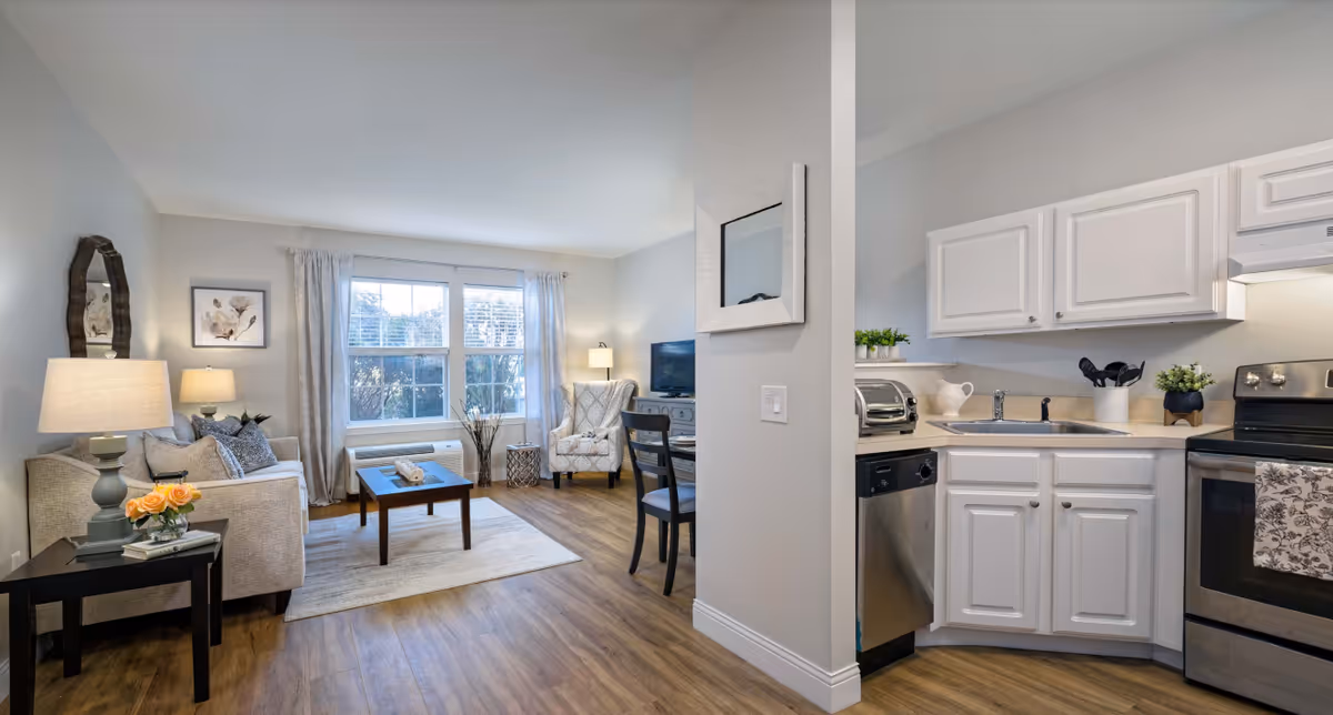 A bright and cozy living area with a beige sofa, a side table with a lamp and flowers, a coffee table on a rug, an armchair near a window with curtains, and a small dining table with chairs. Adjacent to the living area is a compact kitchen with white cabinets, a stainless steel dishwasher, stove, and various kitchen utensils and plants on the countertop.