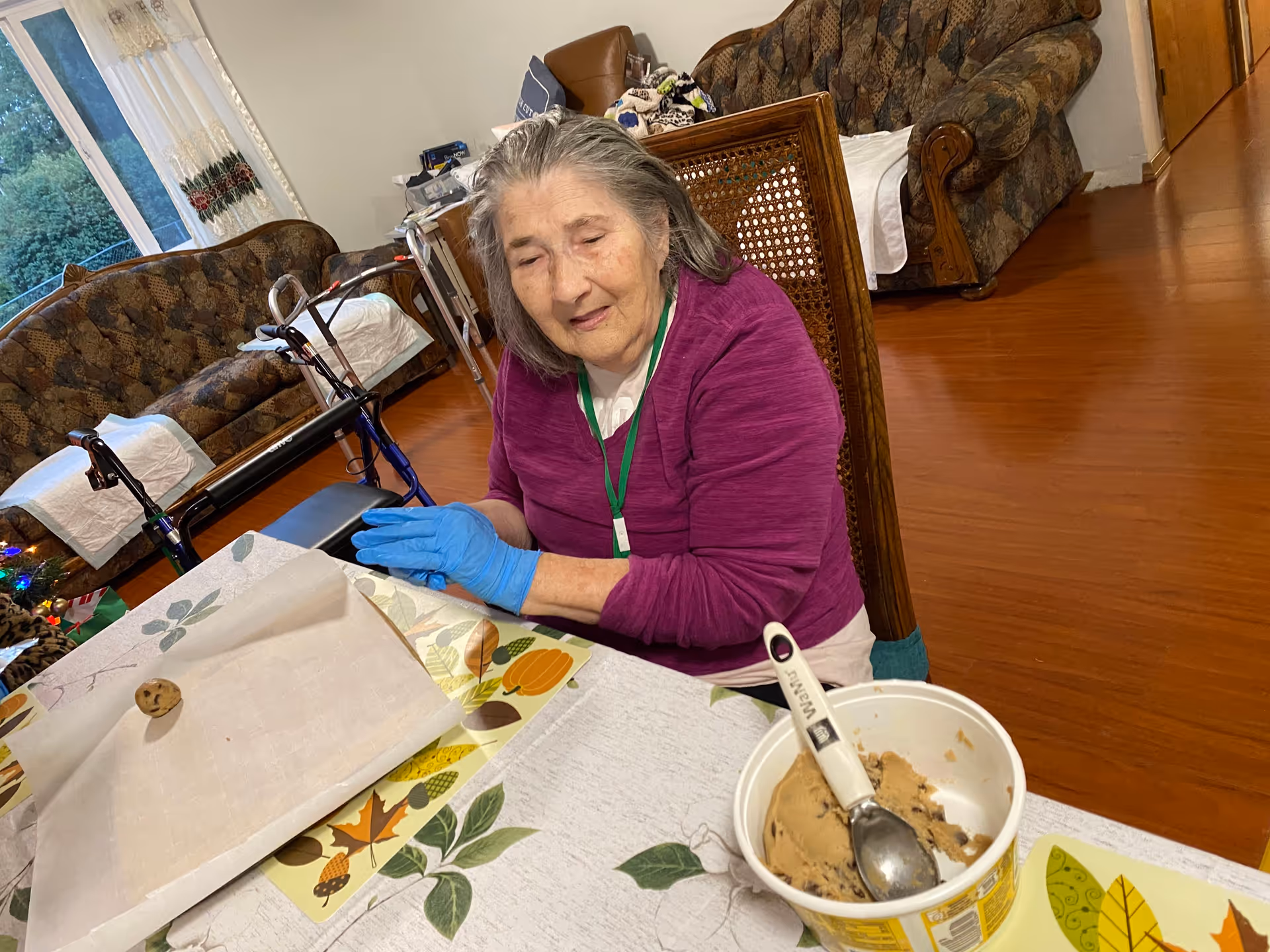 An elderly woman wearing a purple sweater and blue gloves is sitting at a table in a living room. On the table, there is a container of cookie dough with a scoop inside and a baking sheet with one cookie dough ball. The room has wooden floors, patterned sofas, and a window with curtains.