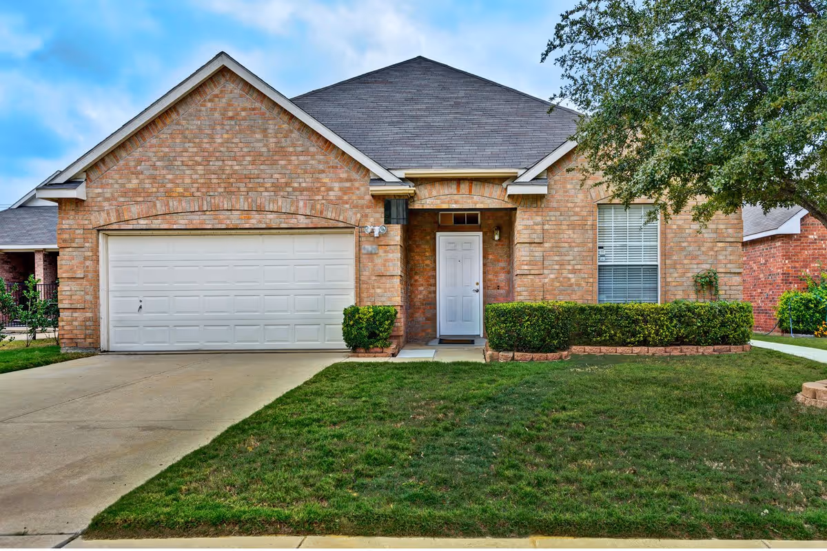 Front exterior view of a single-story brick house with a white garage door, a white front door, trimmed bushes, a tree on the right side, and a concrete driveway leading to the garage.