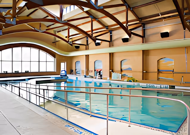Indoor swimming pool with clear blue water, surrounded by a safety railing. The pool area has a high ceiling with exposed wooden beams and large arched windows letting in natural light. The walls are painted in warm tones, and there are poolside chairs and equipment visible.