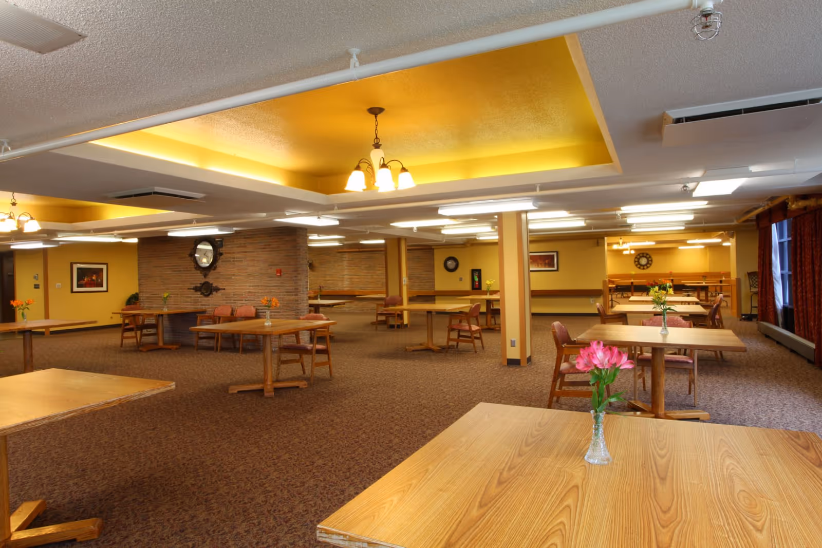 Large communal dining room with wooden tables and chairs, ceiling lighting, and small vases of flowers on the tables.