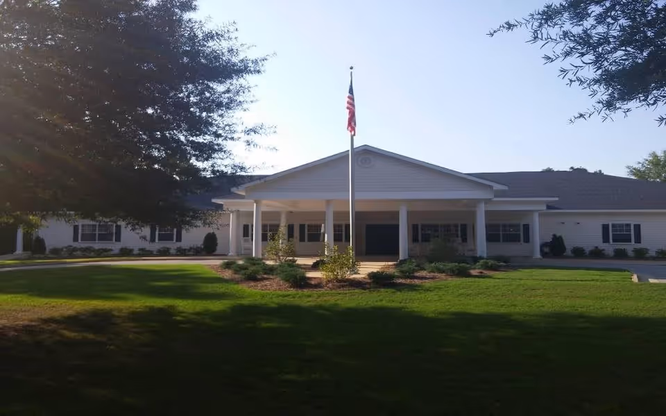 Front exterior view of a single-story building with white siding, a covered entrance supported by columns, an American flag on a flagpole in front, and well-maintained green lawn and shrubs surrounding the area.