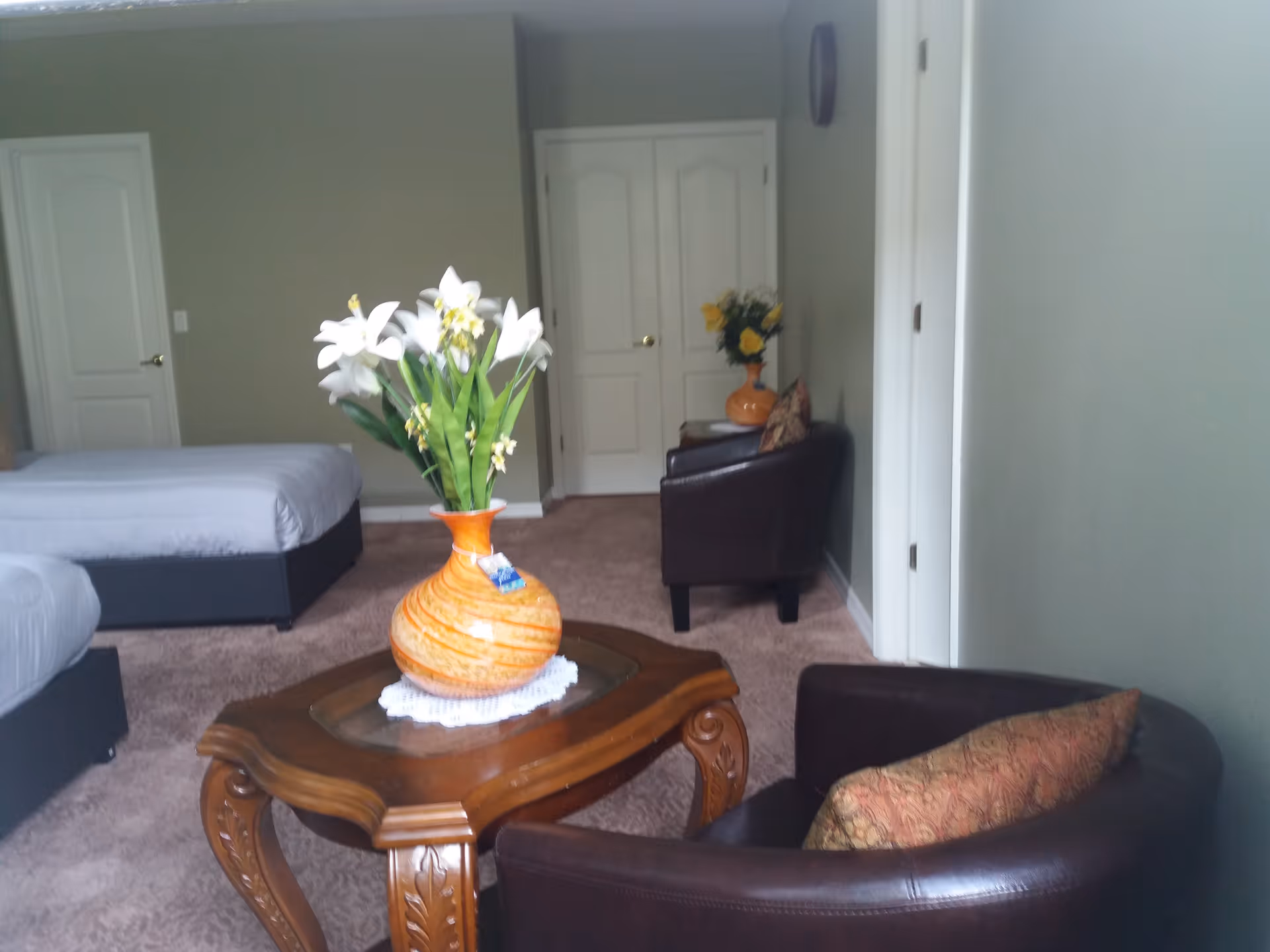 A cozy room with two beds, a wooden coffee table with an orange vase holding white flowers, and two dark brown armchairs with patterned cushions. The room has beige carpet, light green walls, and white doors in the background.