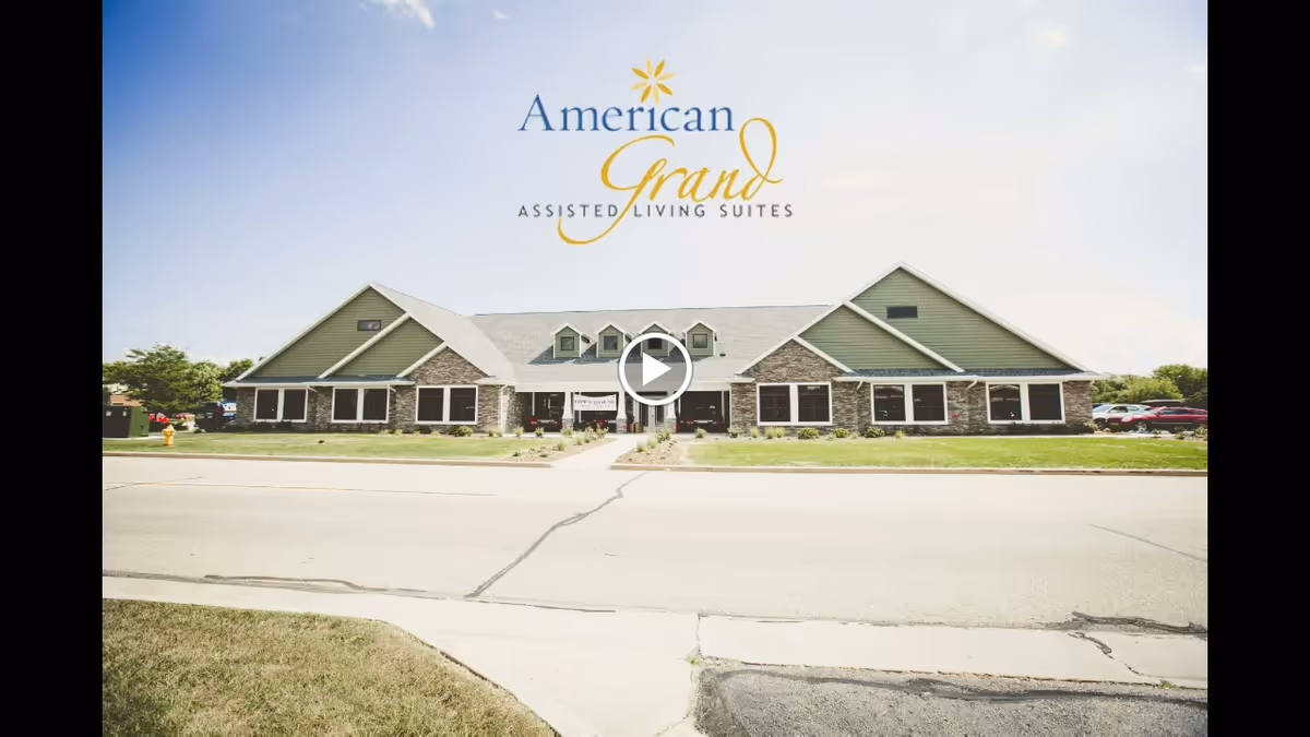 Front exterior view of a single-story assisted living facility building with green siding and stone accents under a clear sky. The building has multiple windows and a central entrance with a pathway leading to the street.