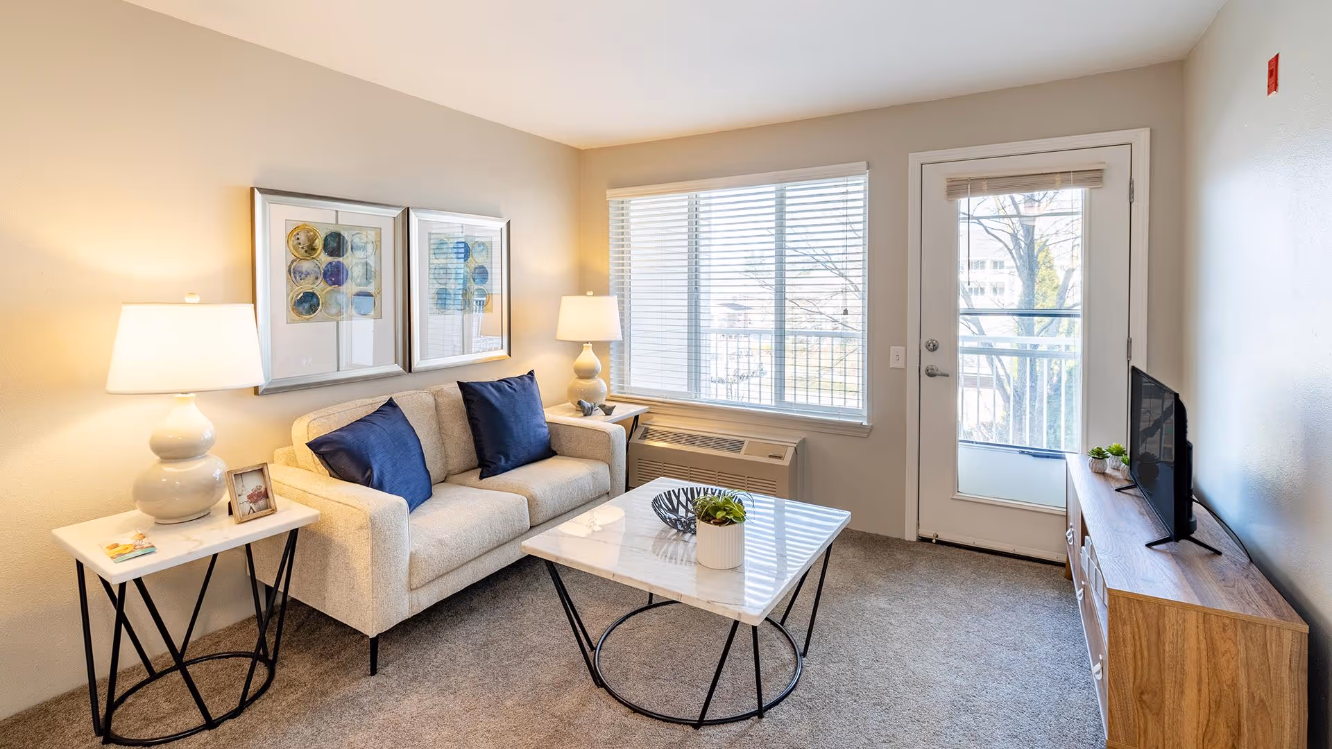 A cozy living room with a beige sofa adorned with two dark blue cushions, a white side table with a lamp and a framed photo, a marble coffee table with a small plant and decorative bowl, a wooden TV stand with a flat-screen TV and small plants, a large window with blinds, and a glass door leading to an outdoor balcony.