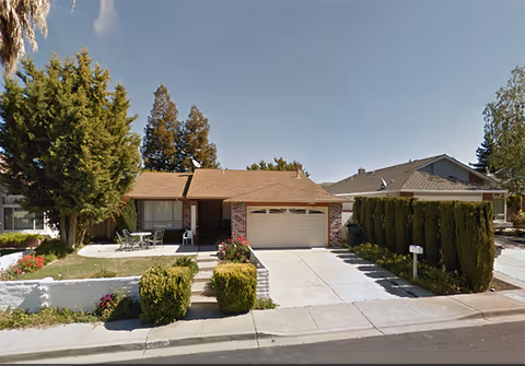 Single-story residential house with a brown shingle roof, a two-car garage, and a front yard featuring trimmed bushes, a small patio with outdoor furniture, and tall trees in the background under a clear blue sky.