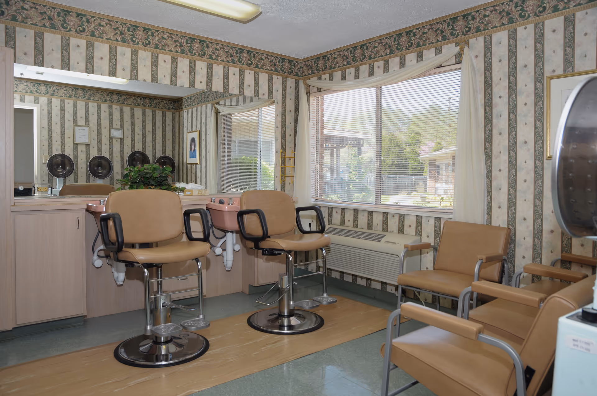 Interior of a salon room with two beige salon chairs in front of sinks and mirrors, additional beige chairs along the wall, floral and striped wallpaper, a large window with blinds, and an air conditioning unit below the window.