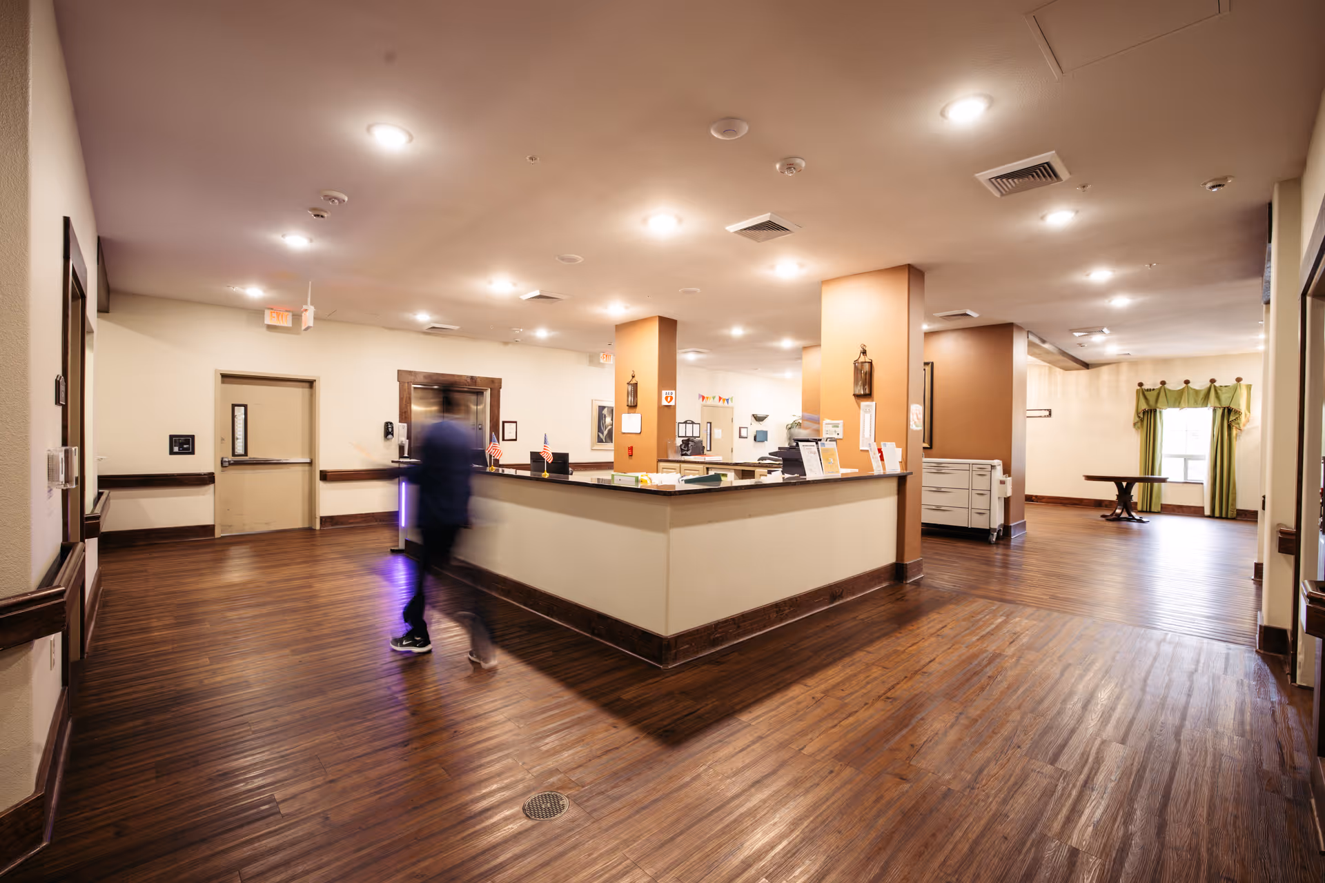 Interior view of a nursing and rehabilitation center reception area with wooden flooring, a central reception desk, warm lighting, and a person walking past the desk. The space has beige walls, some framed pictures, and a window with green curtains in the background.