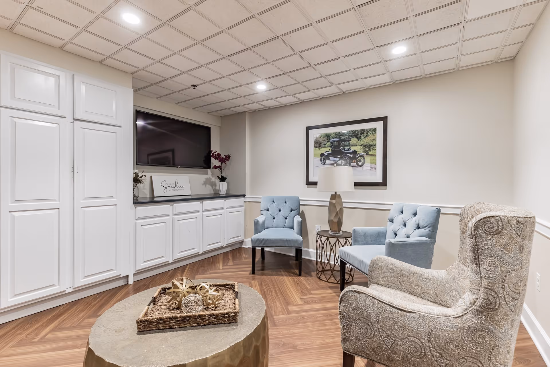 A cozy sitting area in a senior living facility featuring two light blue upholstered chairs, one patterned armchair, a round side table with a lamp, a large flat-screen TV mounted above white cabinetry, and a framed picture of a vintage car on the wall. The room has wood flooring and a tiled ceiling.