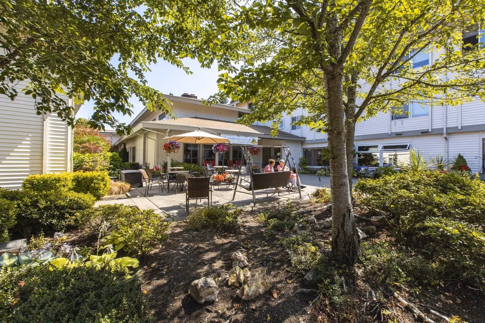 Outdoor patio area at a retirement home with a tree in the foreground, surrounded by bushes and plants. The patio has chairs, tables, a swing bench, and an umbrella. Two elderly people are sitting on the swing bench. The building of the retirement home is visible in the background under a clear sky.