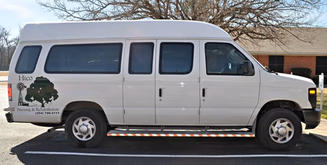 White passenger van with a 'Hico Nursing & Rehabilitation' logo parked outside a brick building.