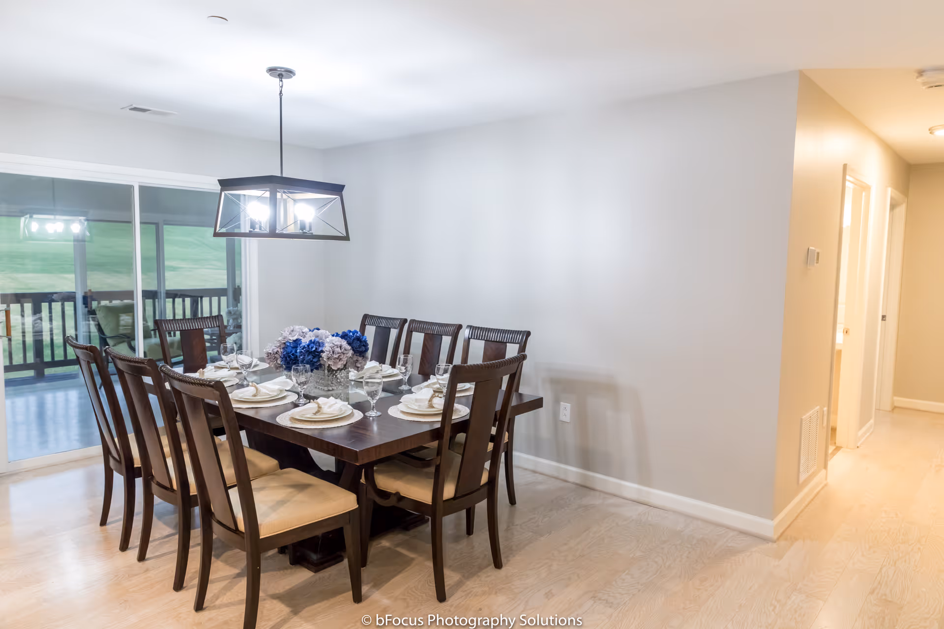 A dining room with a dark wooden table set for eight people with white plates, napkins, and glassware. A centerpiece of blue and white flowers is on the table. The room has light-colored walls and flooring, a modern rectangular chandelier hanging above the table, and sliding glass doors leading to an outdoor patio with a view of a green lawn.