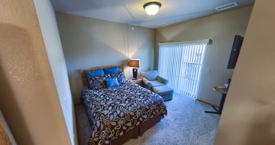 Furnished bedroom with a patterned bed, nightstand and lamp, an armchair with ottoman, and a sliding glass door with vertical blinds.