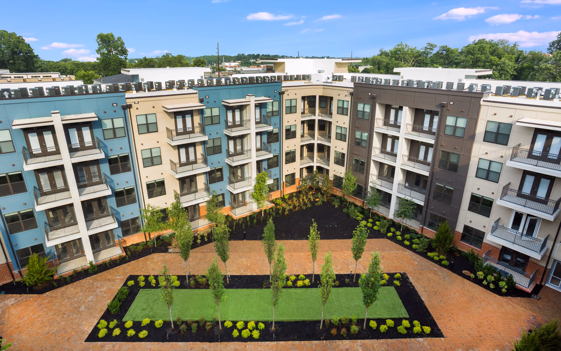 View of a modern multi-story residential building complex with balconies overlooking a landscaped courtyard featuring young trees, shrubs, and brick walkways under a blue sky with scattered clouds.