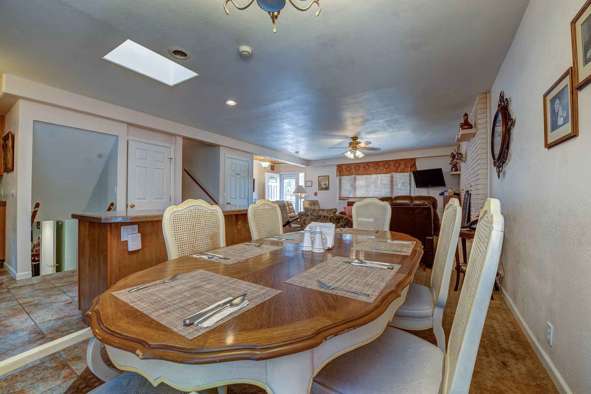 Interior view of a senior living facility dining area with a wooden dining table set for six with placemats and silverware. The room extends into a living area with sofas, a ceiling fan, a wall-mounted TV, and framed pictures on the walls. The floor is tiled near the dining area and carpeted in the living area. There is a skylight and recessed lighting in the ceiling.