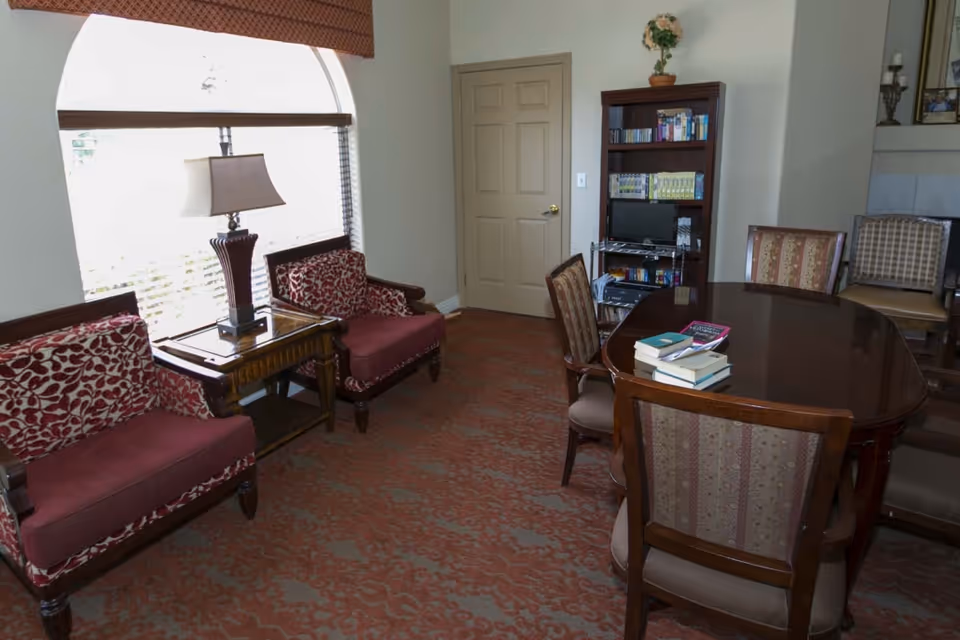 A furnished common room with upholstered chairs, a wooden table with books, a lamp by a large window, and a bookshelf.
