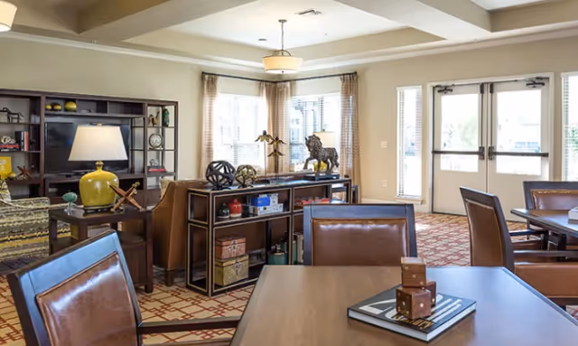 A well-lit common area in a senior living facility featuring a seating area with brown leather chairs and a wooden table with decorative dice, a console table with various decorative items including a lion statue, and a large window with curtains allowing natural light to fill the room. There is a bookshelf with a lamp and other decor, and double glass doors leading outside.
