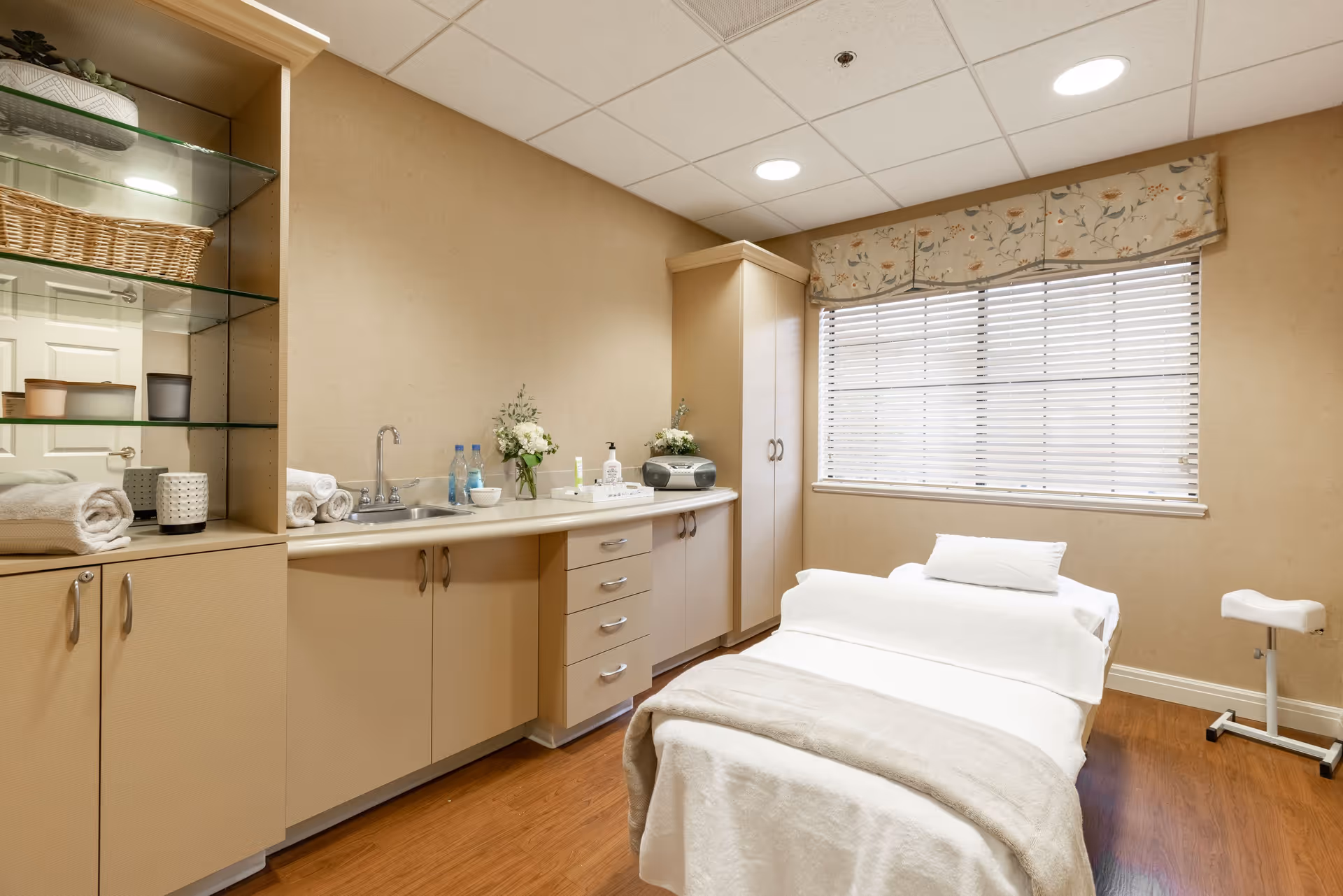 A clean and well-lit room with a treatment or massage table covered in white linens and a beige blanket. The room features beige walls, a large window with blinds and a floral valance, wooden flooring, and built-in cabinets with a countertop holding towels, flowers, bottles, and a small radio. A small adjustable stool is positioned near the window.