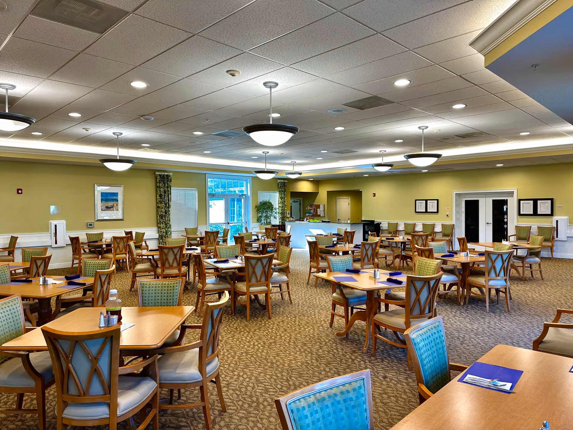 Large, well-lit dining room with multiple wooden tables and chairs arranged for service and place settings on the tables.