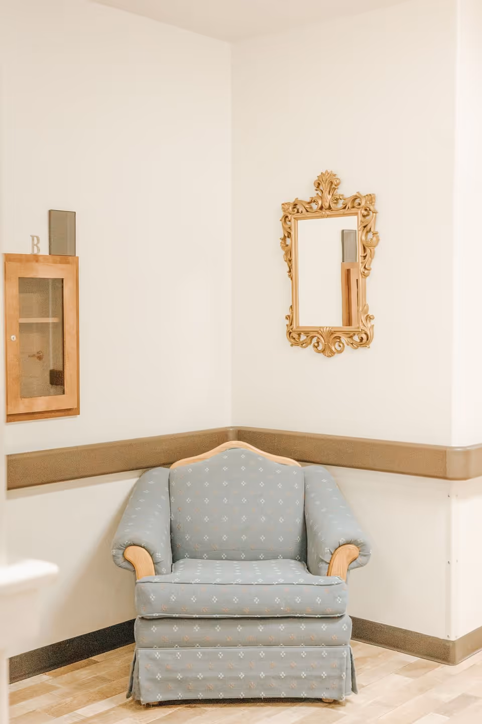 A cozy corner with a light blue upholstered armchair featuring wooden armrests, positioned against cream-colored walls. Above the chair is an ornate gold-framed mirror, and to the left on the wall is a small wooden cabinet with a glass door.